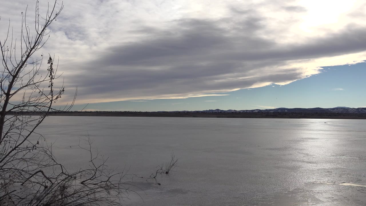 Westminster, Colorado wide view of frozen water with bare branches on the left, a low distant shoreline, snow-dusted hills, gray clouds, sun glare, and no visible people.