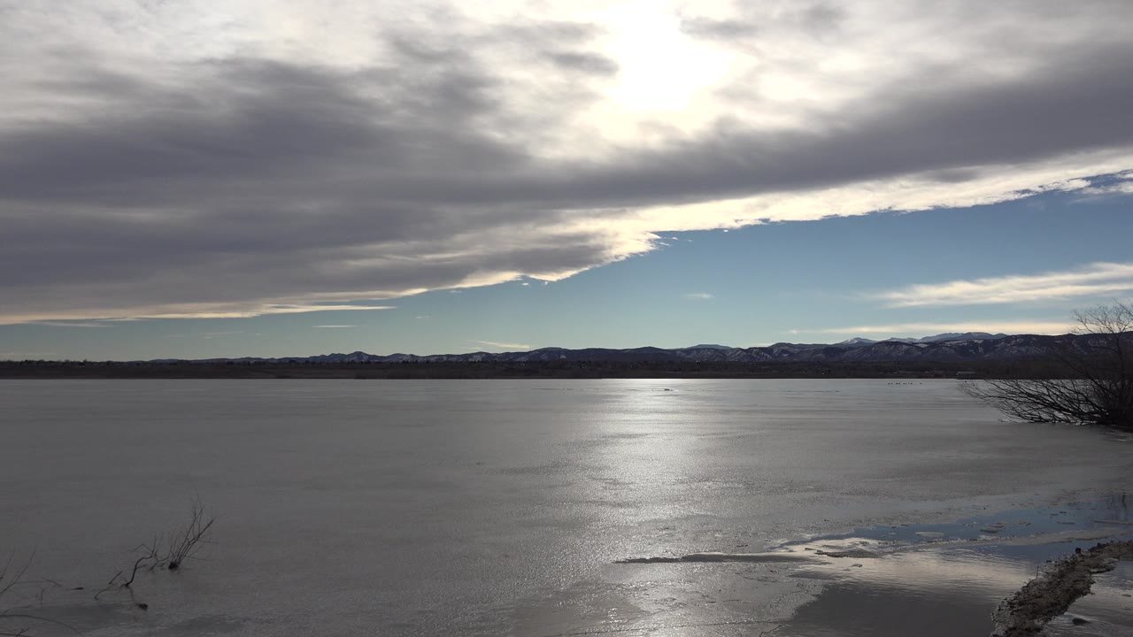 Wide Westminster, Colorado lake view with bright cloud glare over smooth water, distant mountains on the horizon, sparse shoreline brush, and no visible people.