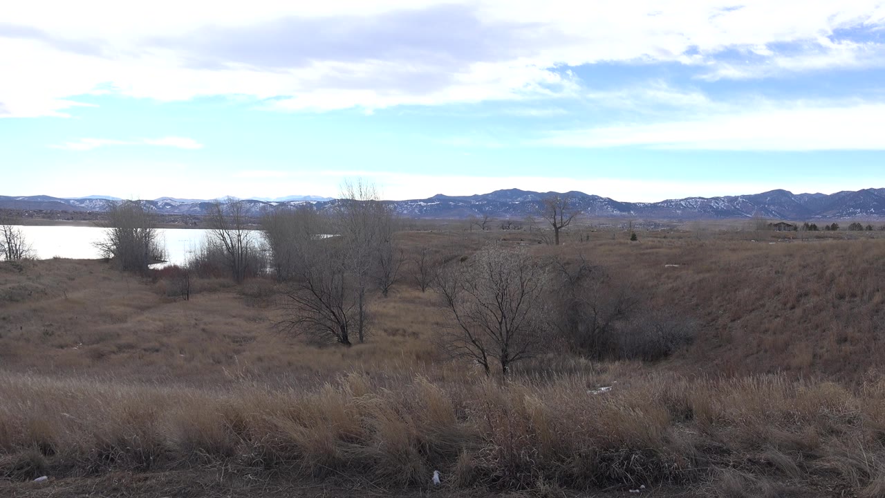 Opening frame of a Westminster, Colorado landscape with dry foreground grass, leafless shrubs near bright water, distant mountains, and no visible people.