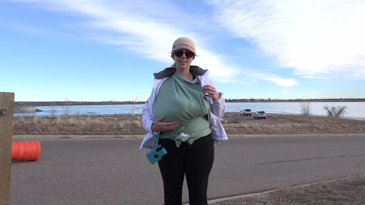 Woman wearing sunglasses and a baby carrier stands by a road near a lake.