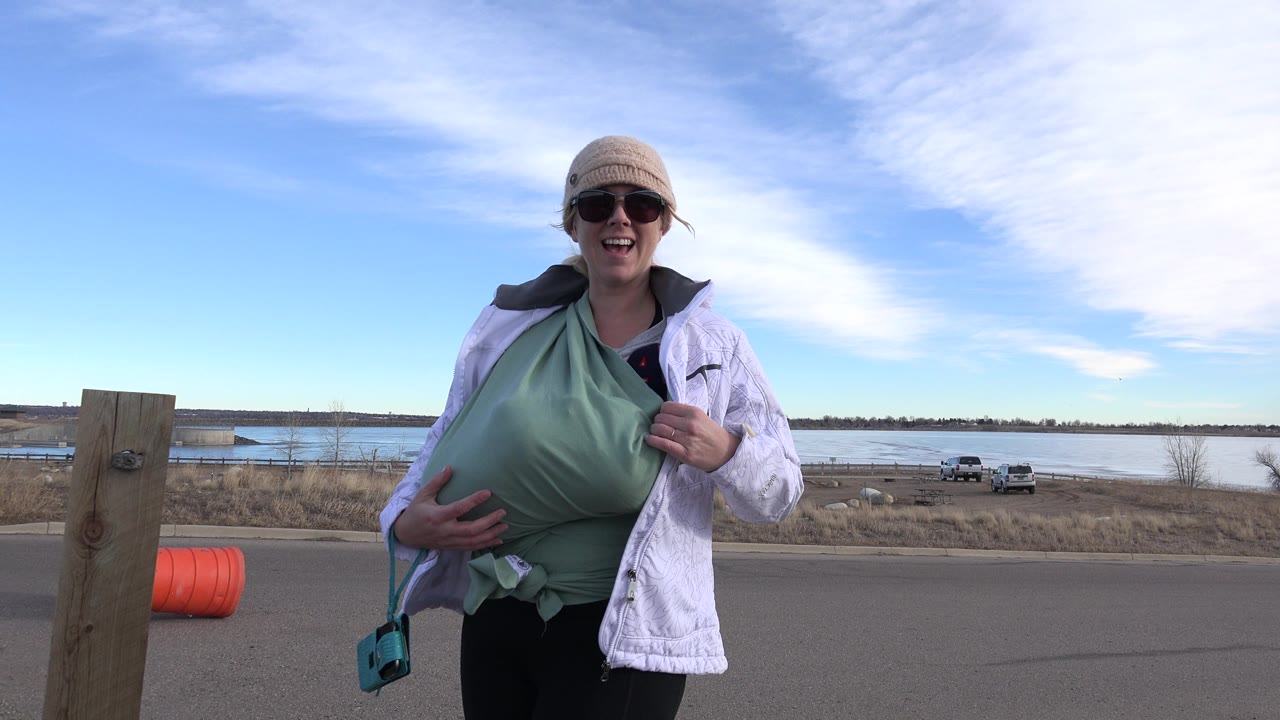 A woman in sunglasses and a knit hat stands beside a paved road near water in Westminster, Colorado, with dry grass, parked vehicles, blue sky, and broad clouds behind her.