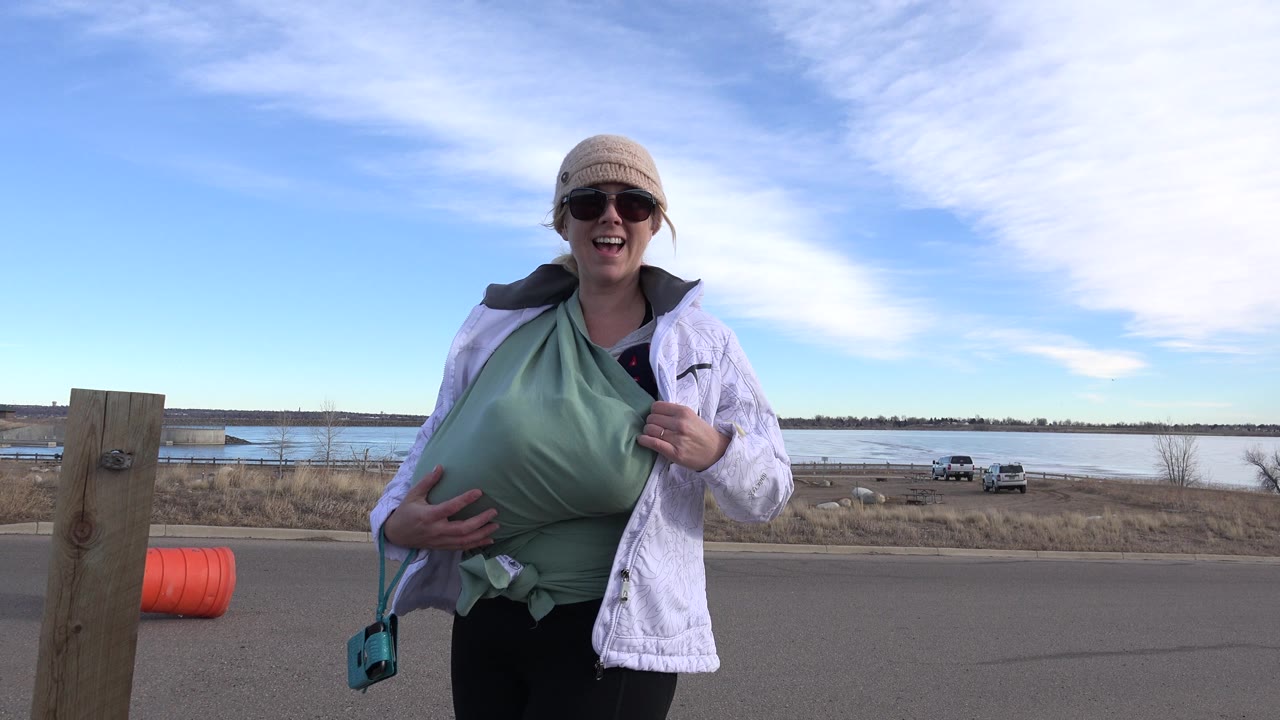 The woman remains near the center of the Westminster, Colorado roadside frame, with a wood post, orange barrel, parked vehicles, water, and high white clouds visible around her.