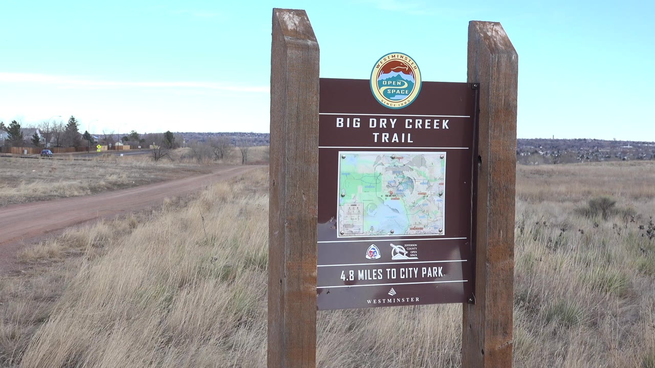 Opening frame of a wooden Big Dry Creek Trail sign in Westminster, Colorado, with a dirt road, dry grass, open field, pale sky, and no visible people.