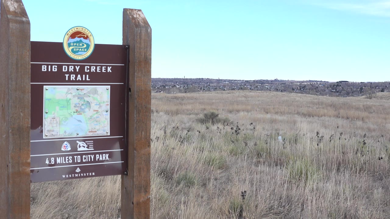 A Big Dry Creek Trail sign and map stand at the left edge of a Westminster, Colorado grassland view, with dry grasses, distant low hills, and no visible people.