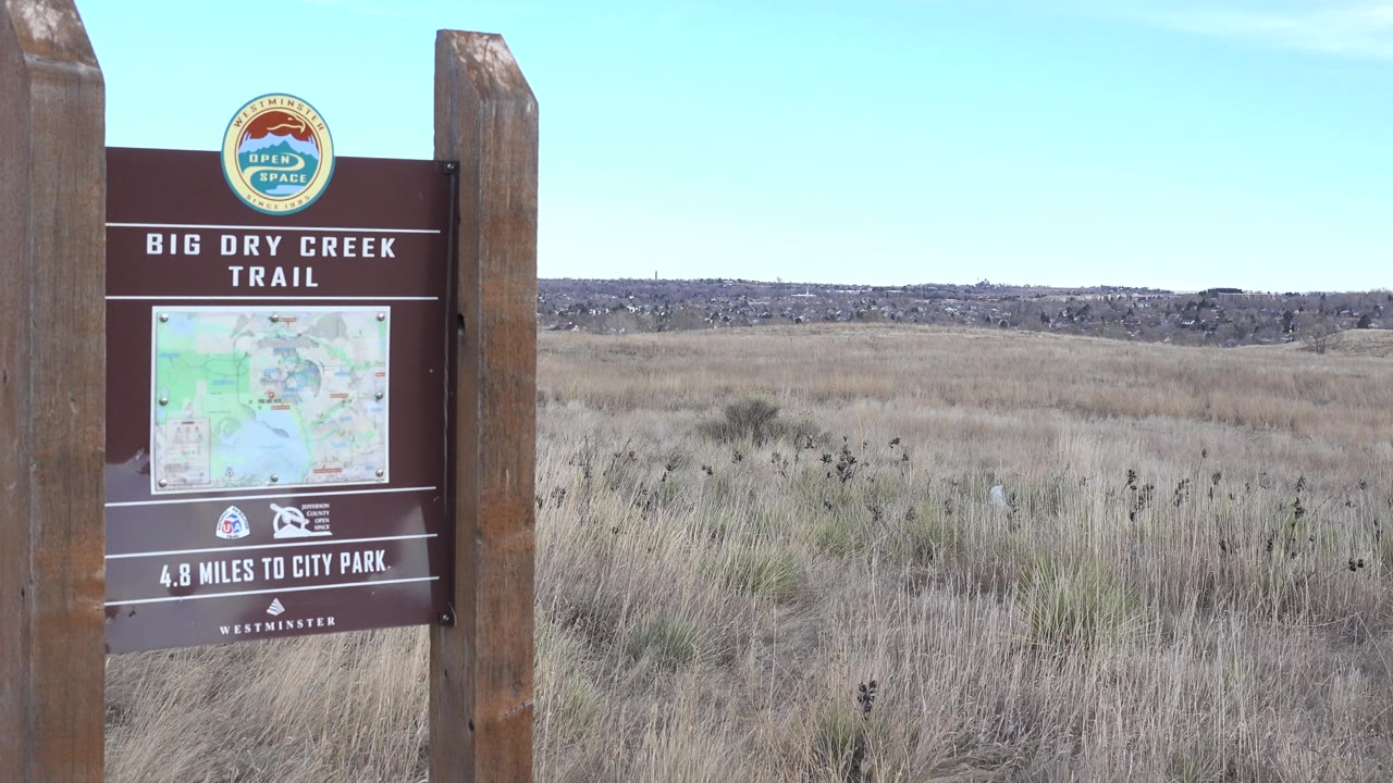 The Westminster trail sign stays near the left side of frame while tan grasses and a low distant ridge fill the background, with no visible people.