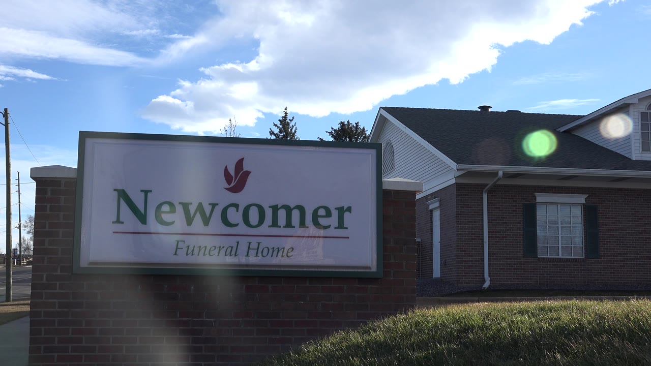 Newcomer Funeral Home sign fills the left side of the frame in Westminster, Colorado, with a brick building, grass, clouds, sun flare, and no visible people.