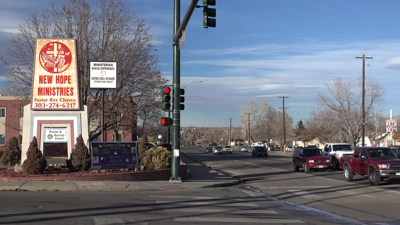 A Westminster, Colorado street corner shows a tall New Hope Ministries sign, red traffic signal, leafless trees, and cars moving through the intersection with no clearly visible pedestrians.