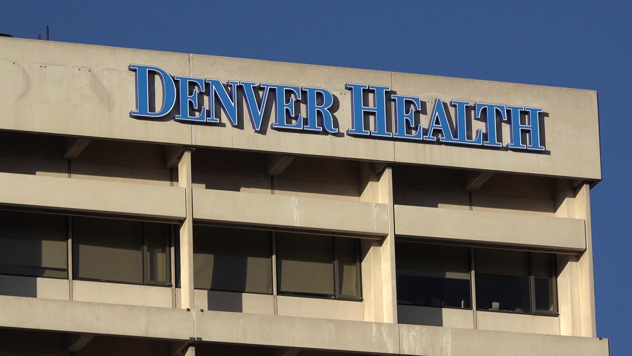 A nearly centered low-angle view holds the blue Denver Health sign above a row of dark windows on the beige building exterior in Westminster, Colorado, with no visible people.