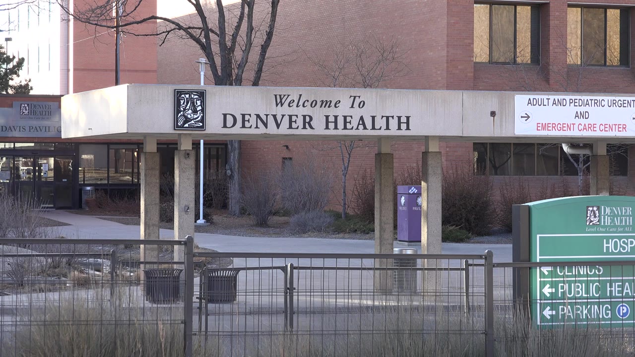 Opening frame of a Westminster, Colorado hospital exterior with Denver Health entrance signage, emergency care direction sign, foreground railings, and no visible people.