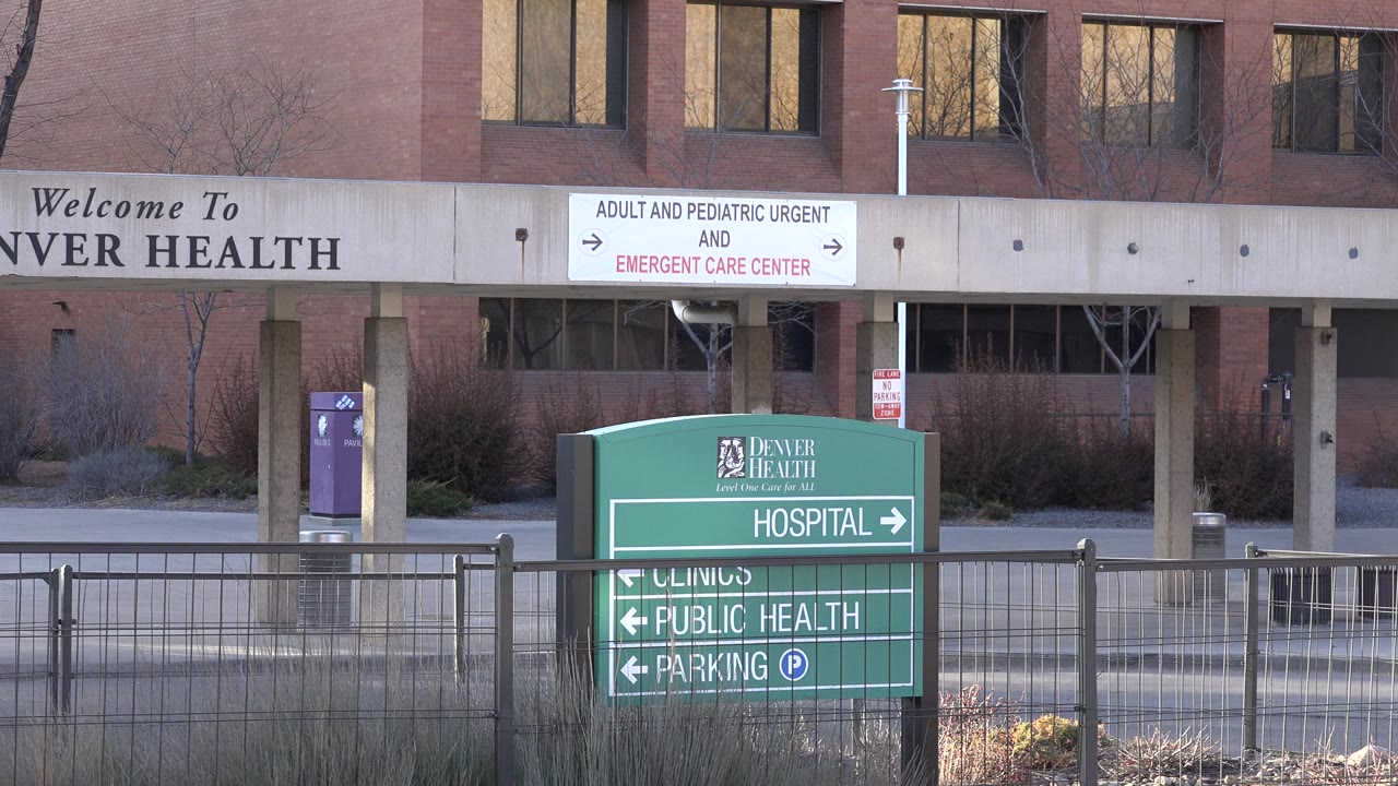 Opening frame of a green Denver Health hospital sign and foreground fence in Westminster, Colorado, with a brick medical building and emergency care signage behind it, no visible people.