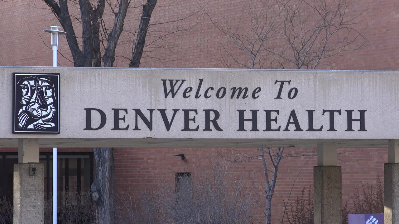 Close frame of the concrete canopy reading Welcome To Denver Health in Westminster, Colorado, with bare trees and brick wall behind it and no visible people.