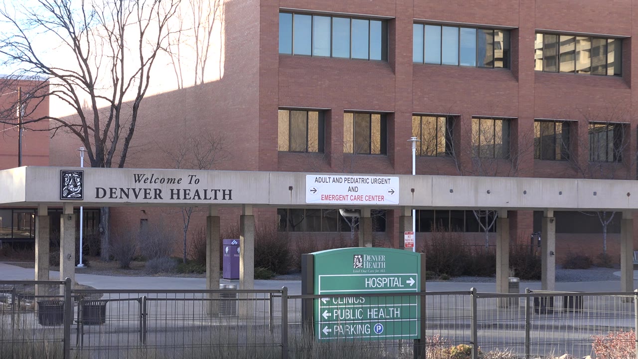Denver Health entrance canopy and green hospital direction sign behind a fence.