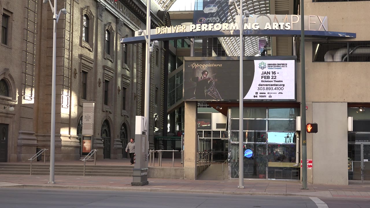 Westminster, Colorado clip frame with the Denver Performing Arts sign above glass doors, a white event board, a dark poster, street fixtures, and a person walking near the left facade.