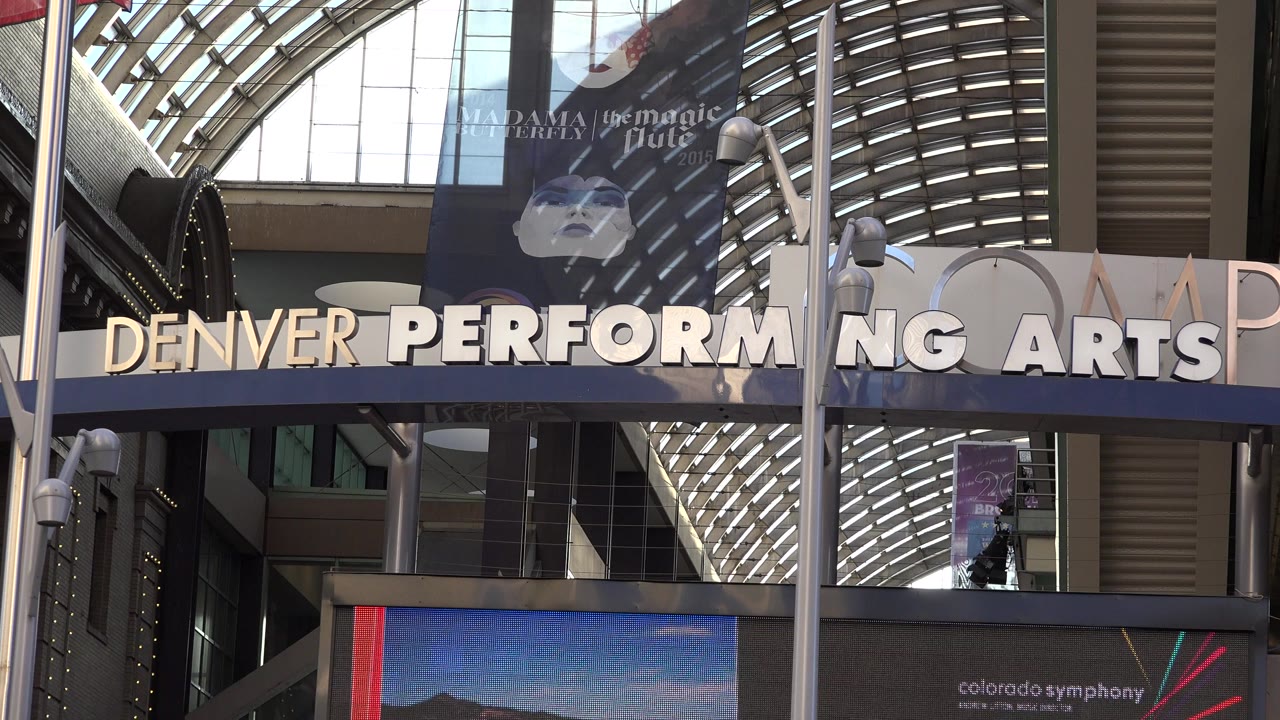 Opening frame of the Denver Performing Arts Center exterior in Westminster, Colorado, with large venue lettering, arched glass roof structure, banners, electronic display panels, and no visible people.