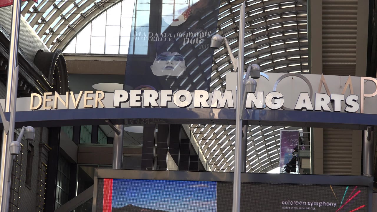 The venue lettering remains centered beneath the glass canopy, with foreground poles cutting through the Denver Performing Arts Center exterior and no visible people.