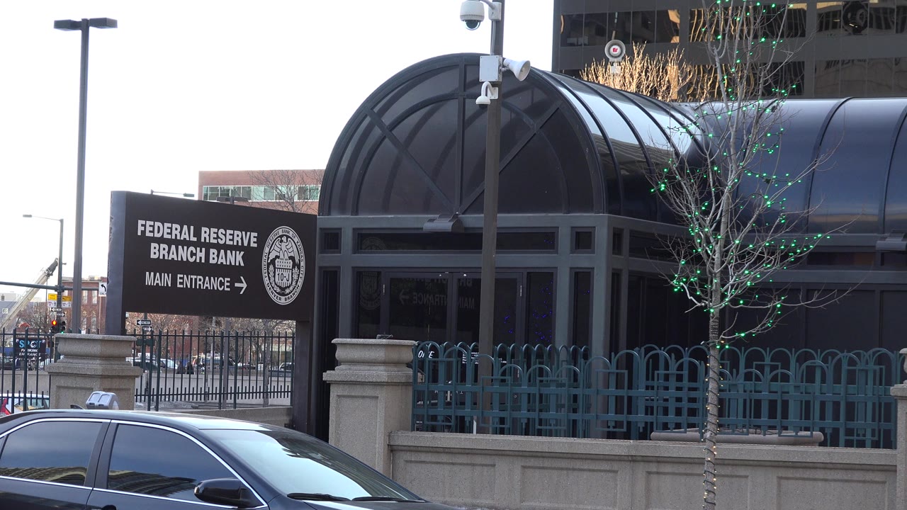 Opening frame of a Westminster, Colorado street with cars in the foreground, a Federal Reserve Branch Bank entrance sign beside a black arched canopy, and no visible people.