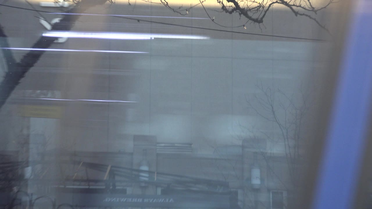 At 0:00 in Westminster, Colorado, the frame is dominated by reflective bus windows showing bare tree branches, building glass, and interior light streaks with no visible people.