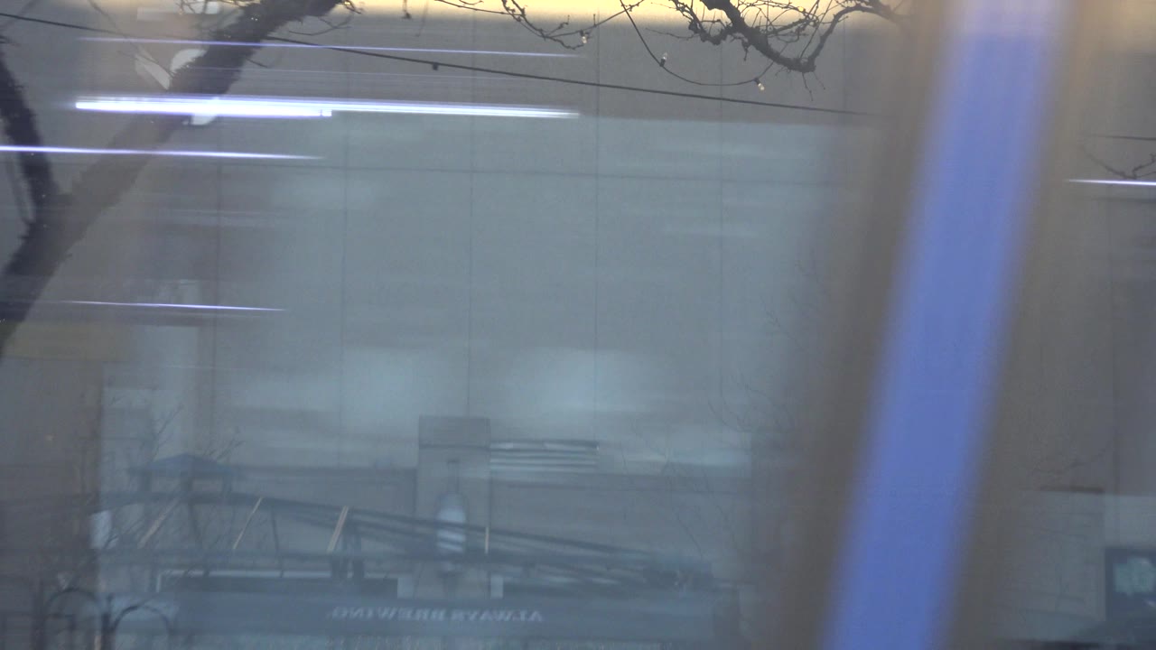 At 0:01 in Westminster, Colorado, the bus still fills the frame, with blue vertical trim, hazy window reflections, bare branches, and a building facade behind the glass, with no visible people.