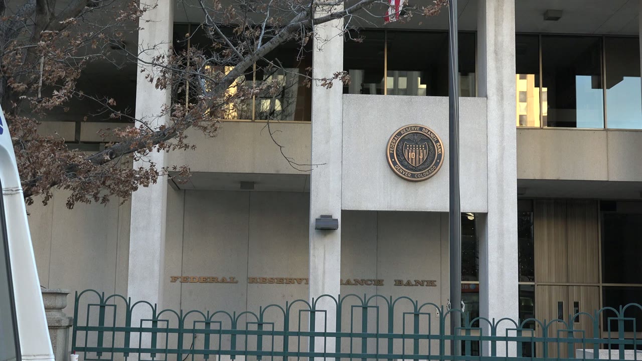 Westminster, Colorado public building at 0:01, with Federal Reserve Bank lettering partly visible across the facade, bare branches in front, green railings, and no visible people.