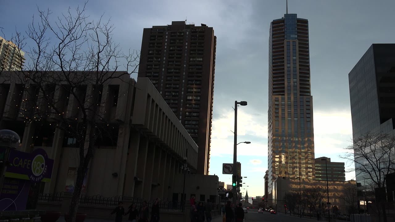 Dark downtown street with tall buildings, bare trees, traffic lights, and pedestrians near the sidewalk.