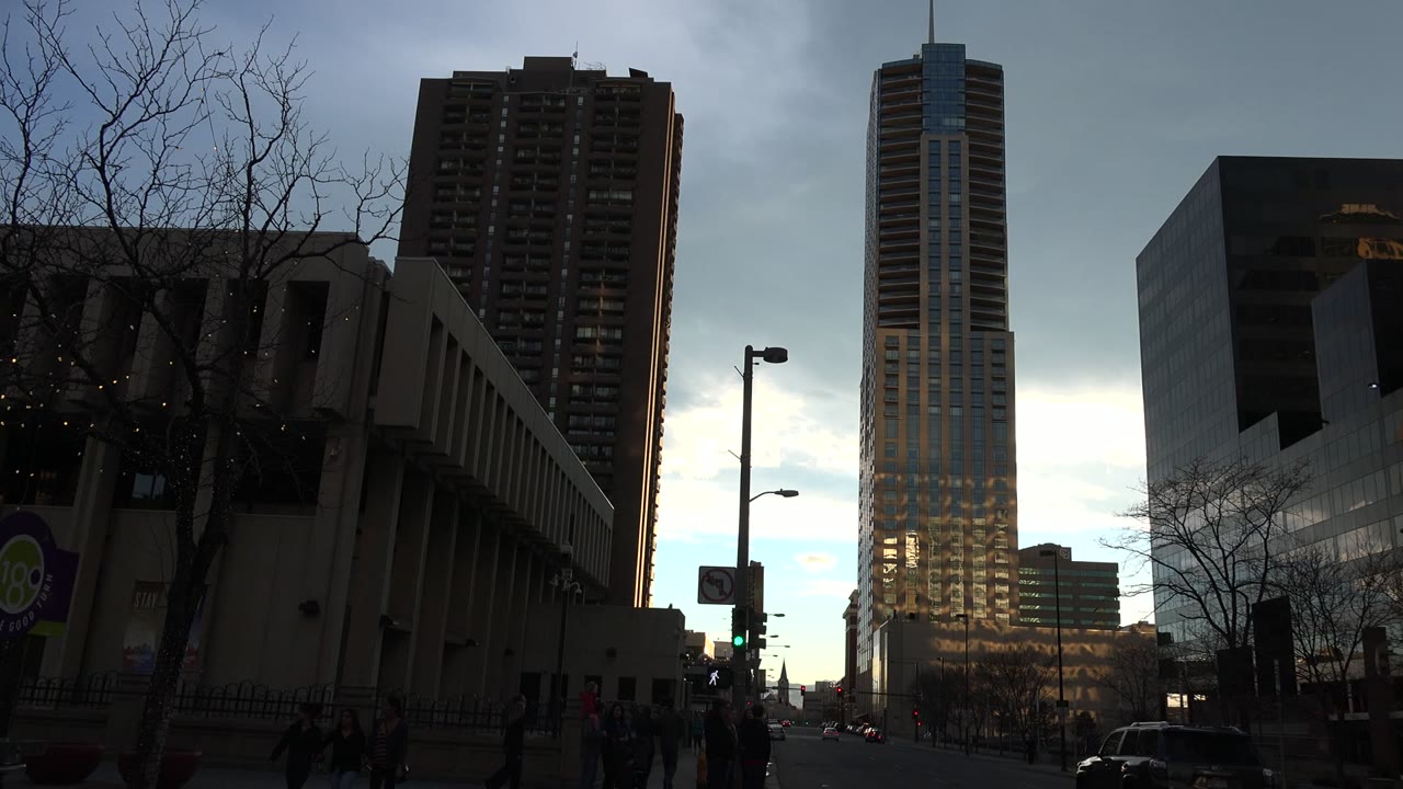 Westminster, Colorado street frame with a reflective glass tower centered in the background, darker buildings on both sides, cars near the intersection, and pedestrians visible below.