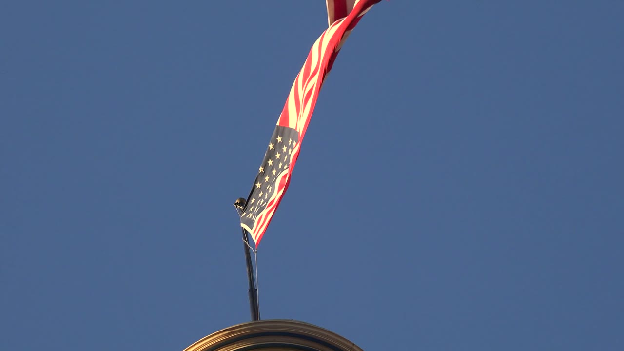 American flag and red-white windsock above a building roof against blue sky.