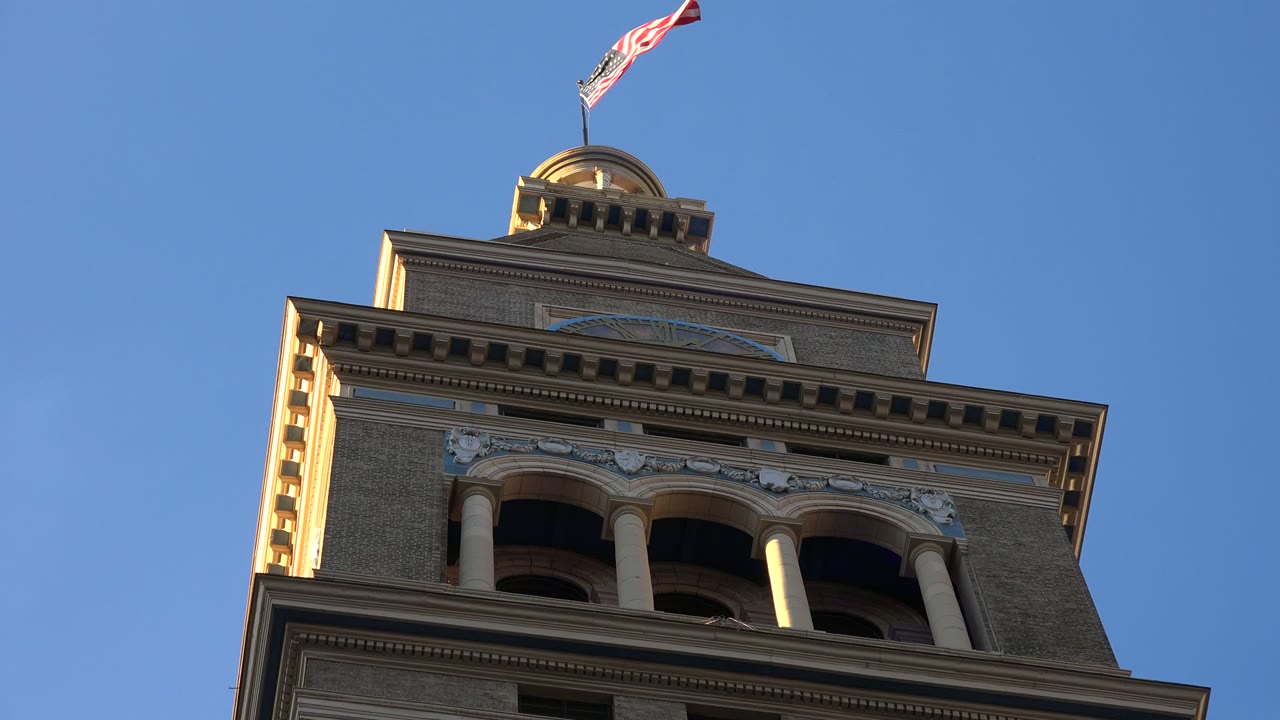 A closer low-angle moment on the Westminster, Colorado tower shows the flag shifted in the wind above the roofline and no visible people in frame.