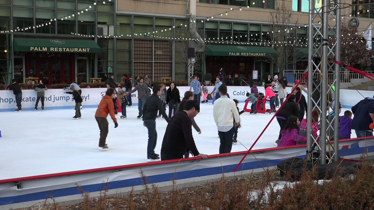 Wide view of people skating on an outdoor ice rink in Westminster, Colorado, with a red rink rail, dried plants, string lights, and Palm Restaurant signage behind them.