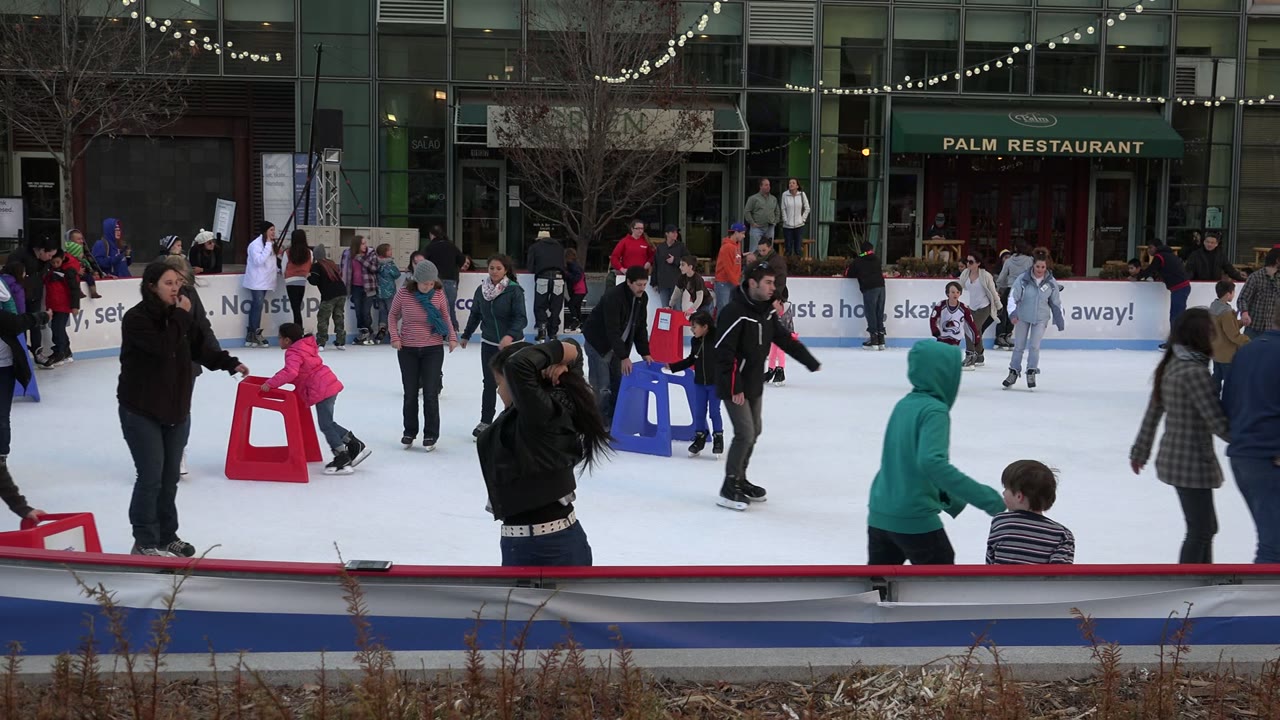 Families skate on an outdoor ice rink near storefront windows.