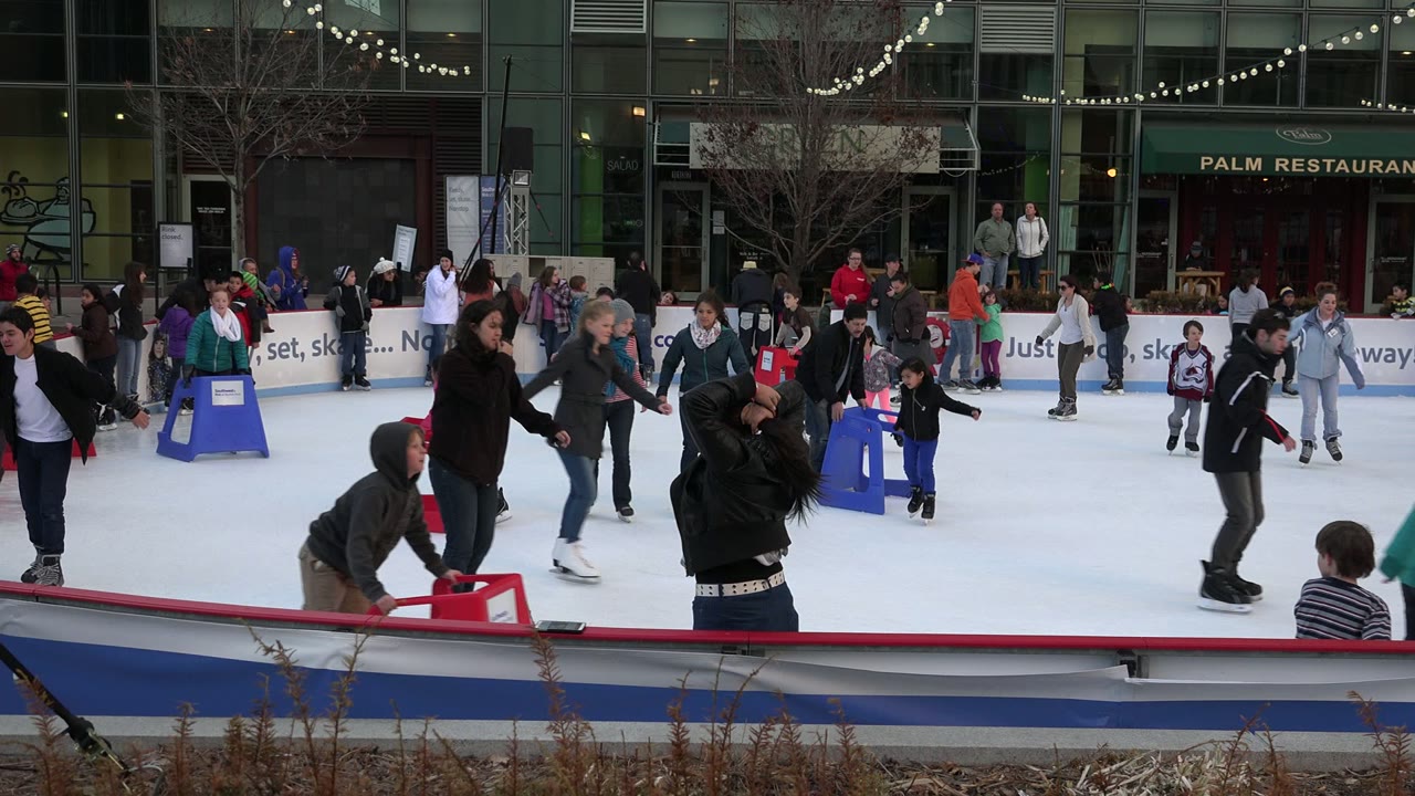 Skaters spread across the Westminster outdoor rink as a child in a green hoodie and nearby adults pass red and blue skating aids beside the rink boards.