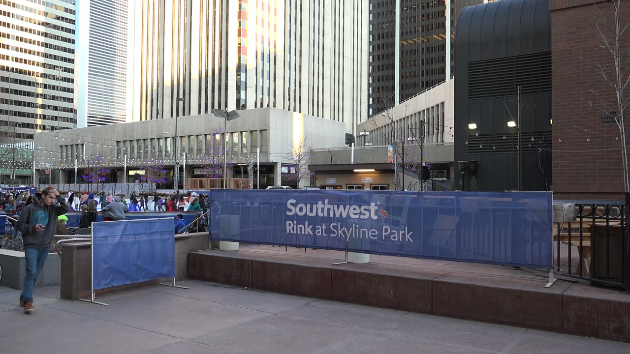 Sidewalk beside a blue rink barrier with pedestrians and tall buildings.