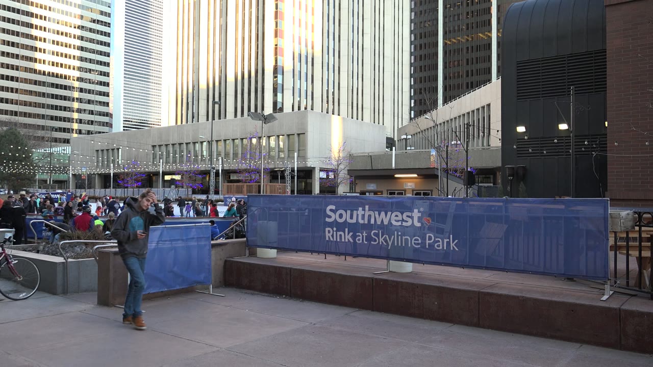A man walks along the Westminster, Colorado sidewalk past the blue rink barrier, with bicycles parked near a planter, people seated or standing beyond the rail, and reflective high-rises in the background.