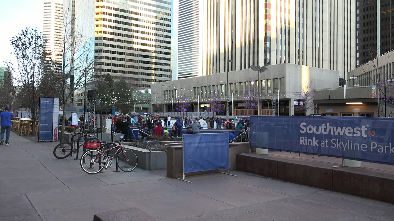 Opening frame of a Westminster, Colorado outdoor ice rink with people on and around the ice, blue Southwest Rink barriers, parked bicycles, bare trees, and tall buildings in warm light.