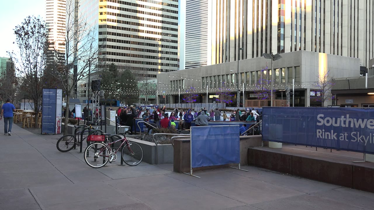 Crowded outdoor skating rink beside blue Southwest Rink barrier and high-rise buildings.