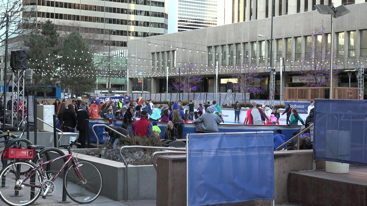 Outdoor ice rink with skaters, spectators, bicycles, and city buildings.
