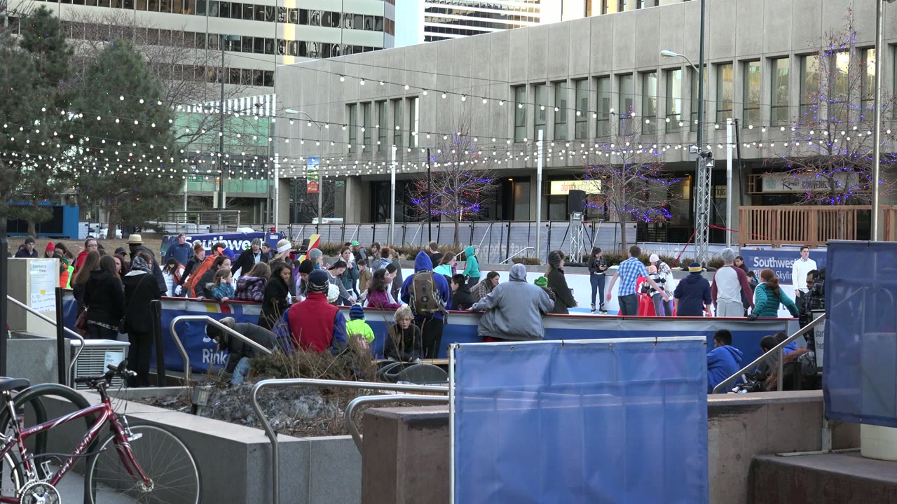 Westminster, Colorado rink scene with a dense group of skaters on the ice, spectators along the barrier, string lights overhead, and office-style buildings in the background.
