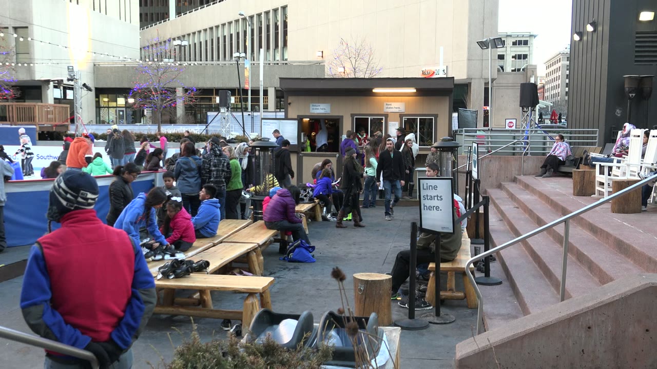 Crowd gathered near benches and a small tan building in an outdoor plaza.