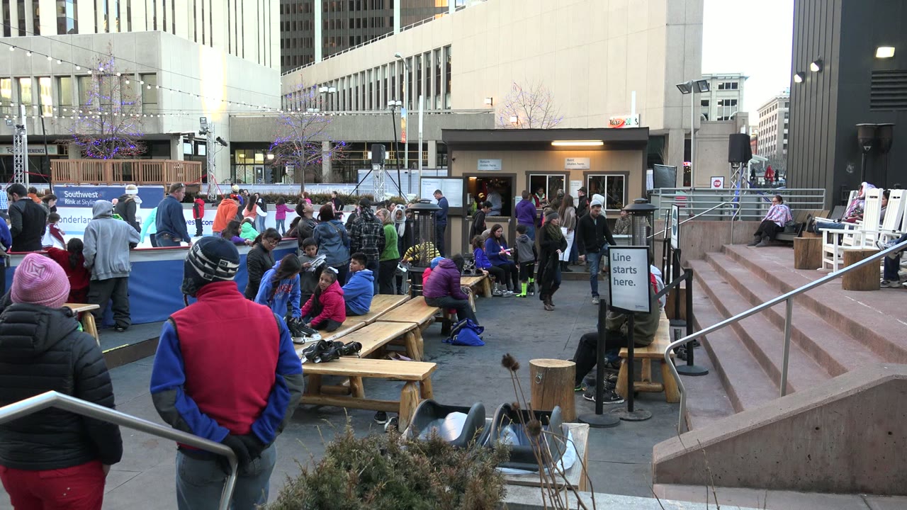 A Westminster, Colorado plaza scene shows people in winter clothing beside benches, with a line sign, blue barrier, and tan building visible behind them.