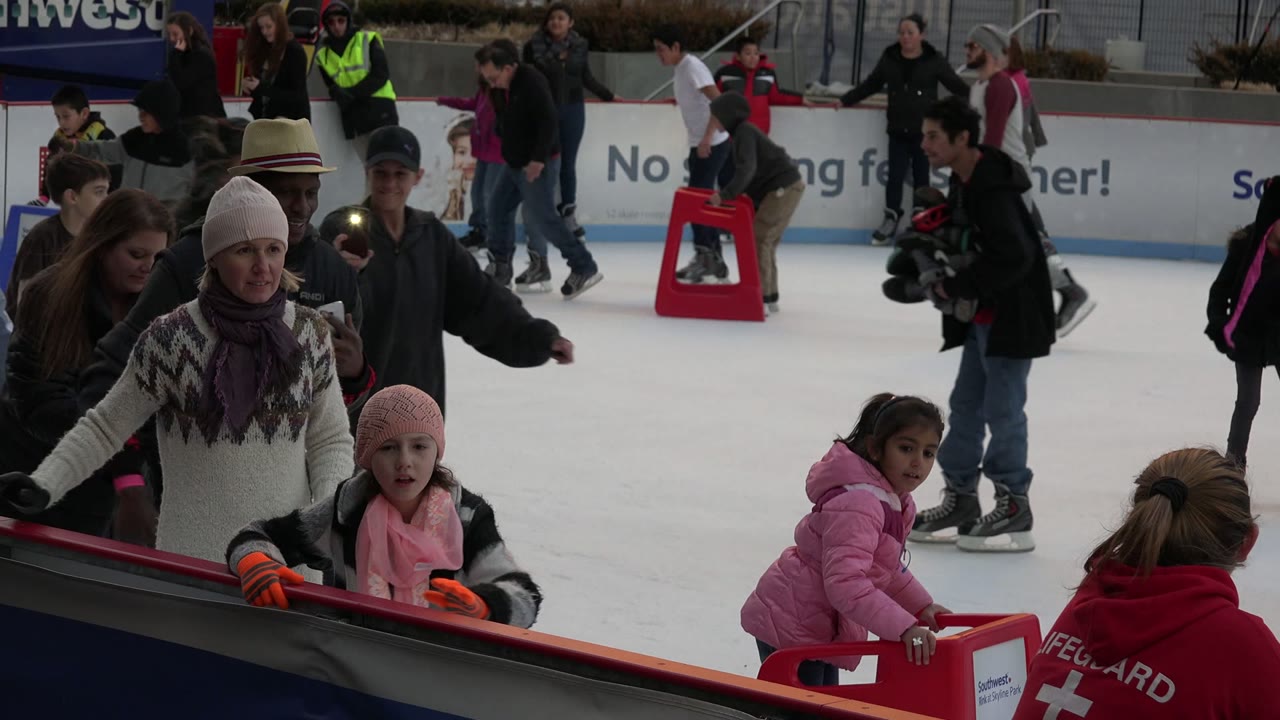 At an outdoor ice rink in Westminster, Colorado, a child in a pink hat stands near the foreground rail while adults and other skaters fill the ice behind her.