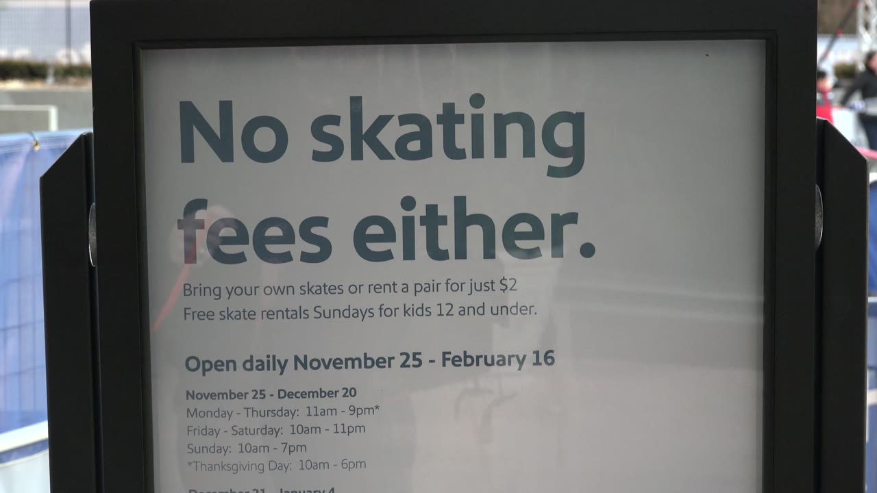 Close foreground view of a No skating fees either sign at an outdoor skating rink in Westminster, Colorado, with rink barriers, purple-lit trees, buildings, and distant people visible behind it.