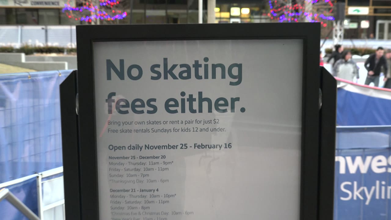 Mid-clip close view of the No skating fees either sign in Westminster, Colorado, with blue rink boards, scattered skaters, string lights, and modern building facades in the background.