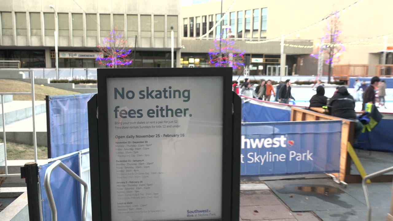 Right-framed No skating fees sign in Westminster, Colorado, with the Southwest at Skyline Park barrier, rink entrance area, skaters, seated viewers, and bright late-day light on nearby buildings.