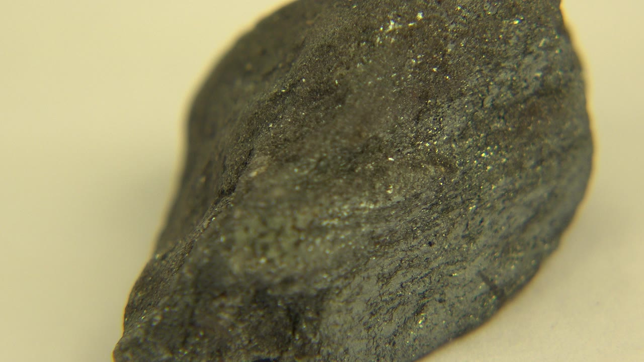 A dark rough rock sits in tight close-up on a cream tabletop in Westminster, Colorado, with faint crystal flecks visible and no visible people.