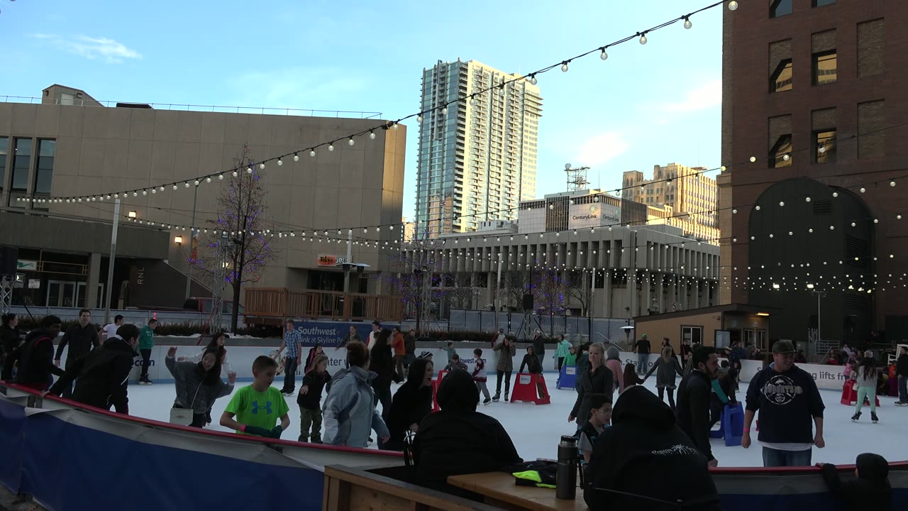 Outdoor ice rink with skaters, string lights, and city buildings.