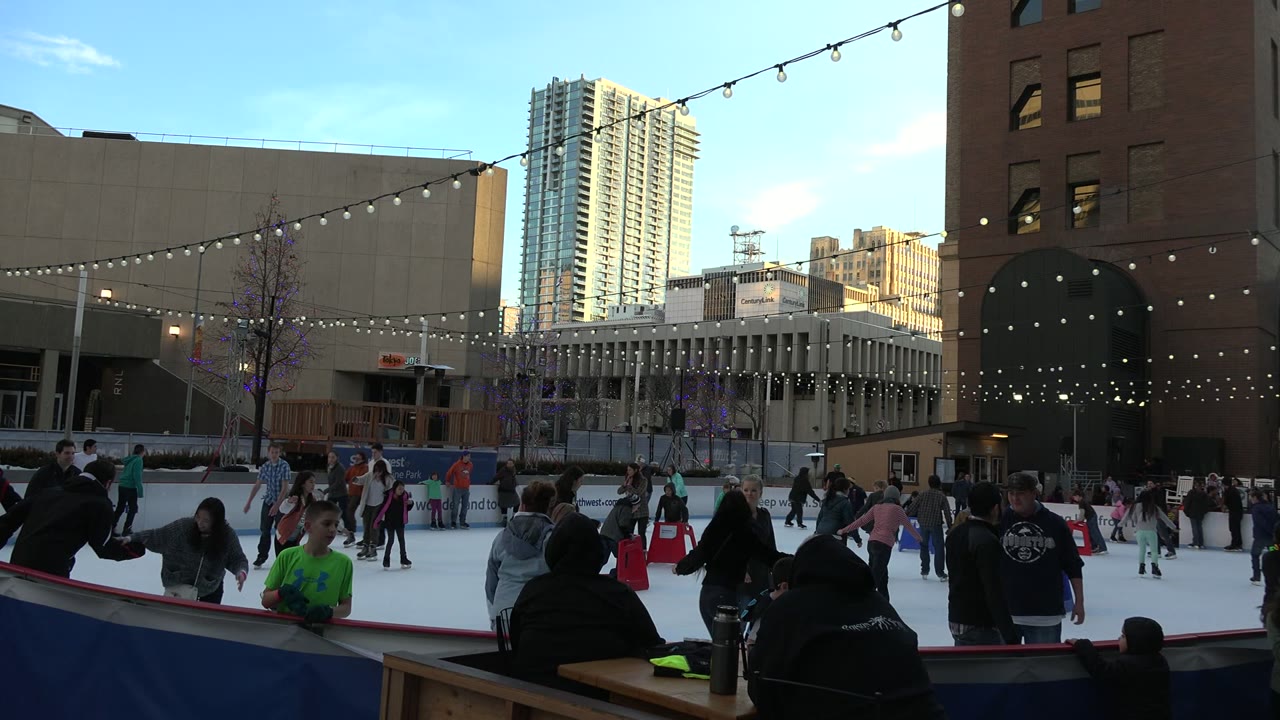 Skaters move across the Westminster outdoor rink while dark foreground figures stand or sit near the boards and high-rise buildings fill the background.