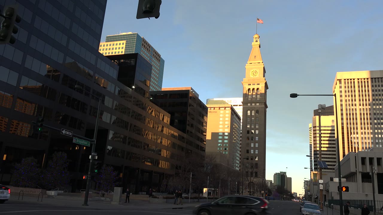Westminster, Colorado urban roadway with dark glass buildings on the left, a sunlit tower near center, a white car in the foreground, and pedestrians visible at sidewalk distance.