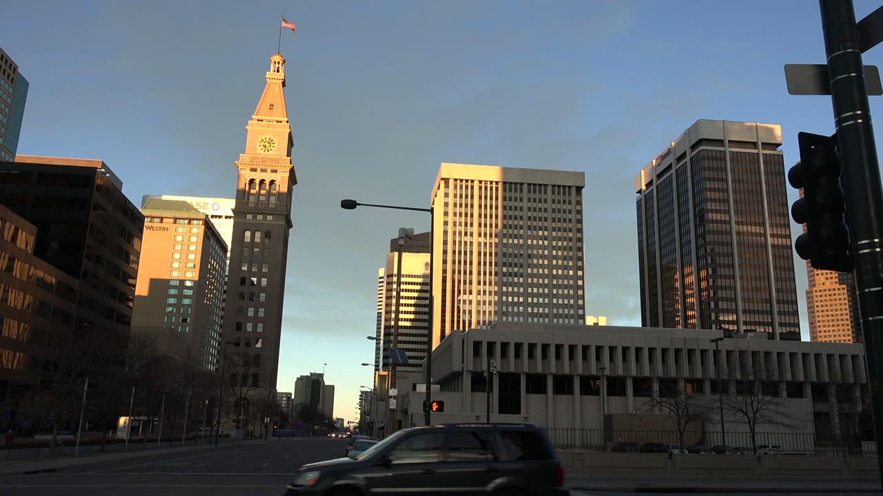 Westminster, Colorado street view with the clock tower near center, bright upper building faces, a traffic signal pole in the foreground, and small distant pedestrians.