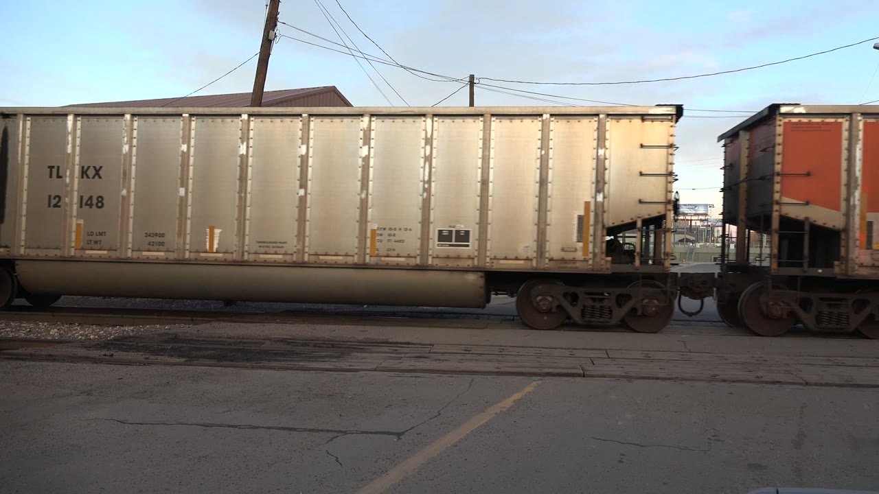 A tan freight rail car fills the frame in Westminster, Colorado, with steel wheels on tracks, cracked pavement in front, utility wires above, and no visible people.