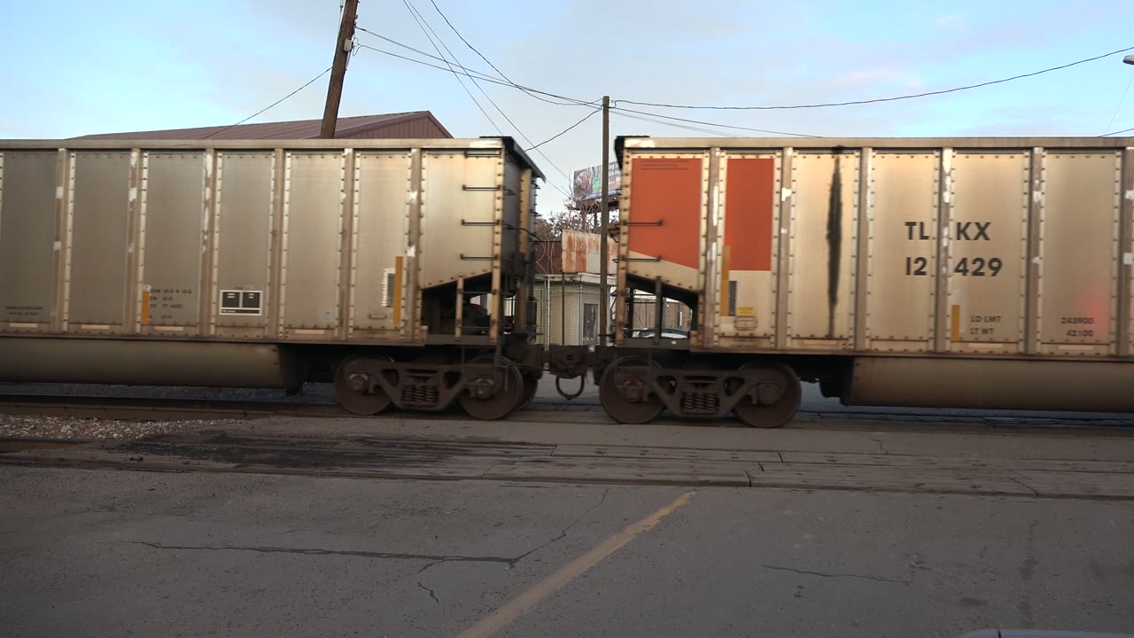 A tan freight car remains centered in Westminster, Colorado, with a red end panel near the left edge, wheel trucks below, a low building behind it, and no visible people.