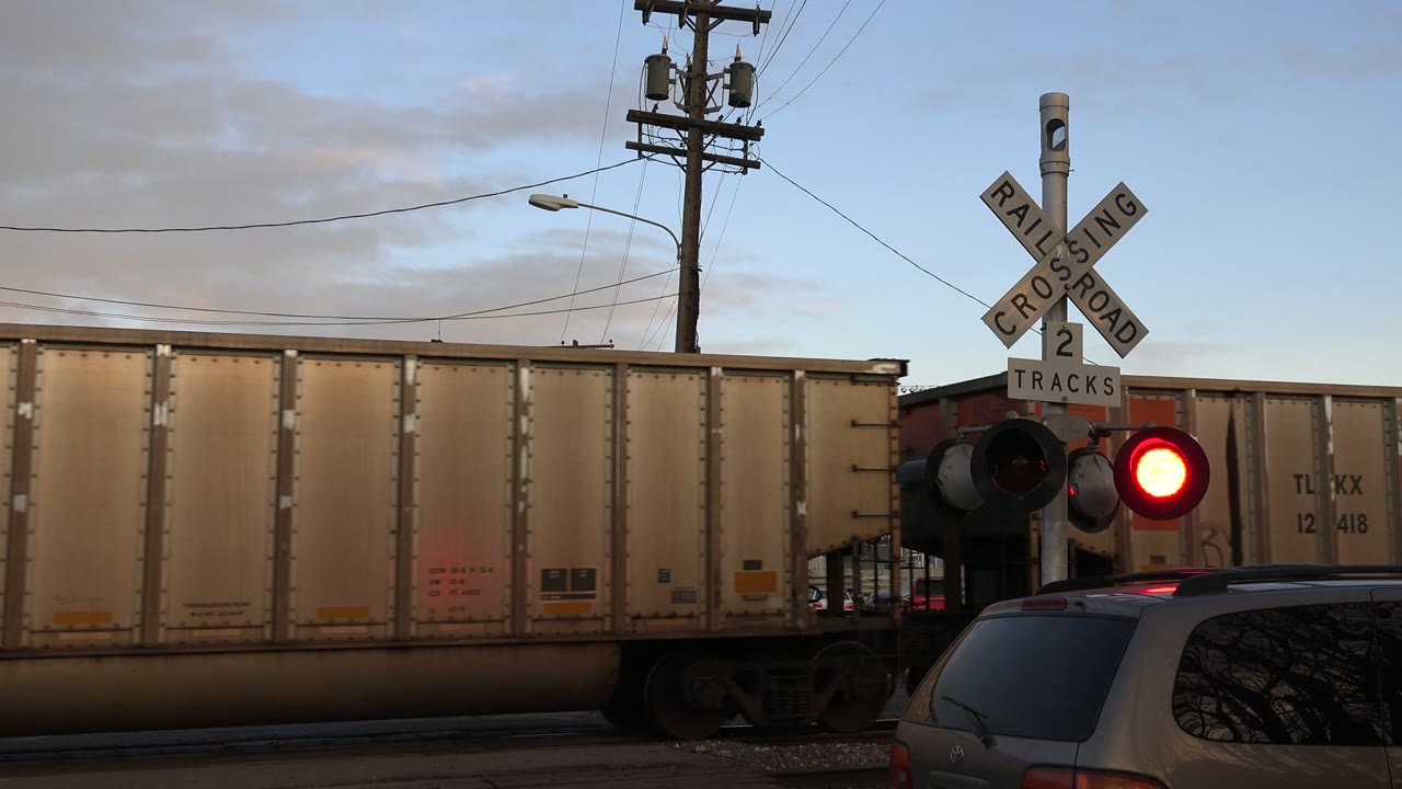 A freight car moves farther across the Westminster crossing while the red signal glows beside a partial vehicle roof in the foreground and no visible people appear.